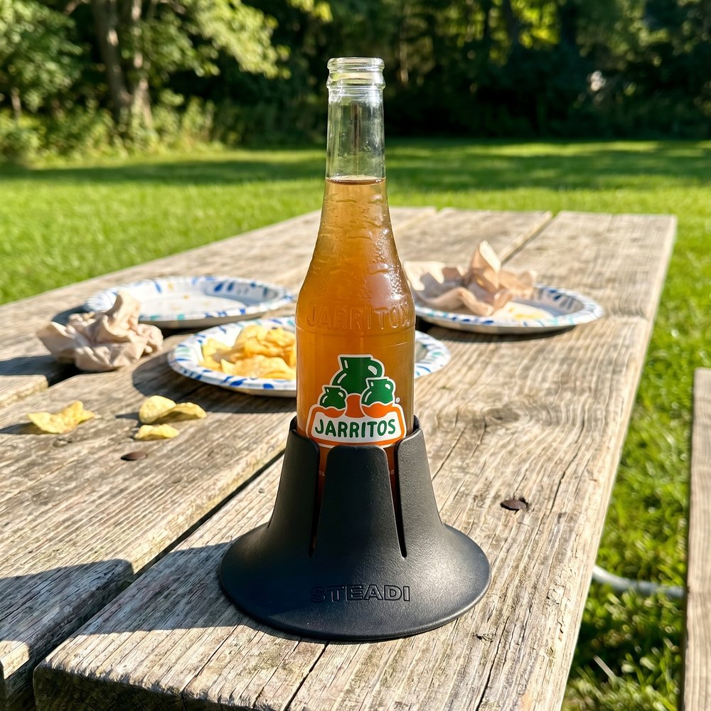 A bottle standing in a black Steadi on a weathered wooden picnic table with food and trees behind.